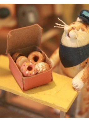 Mixed media narrative sculpture of a cat with a conductor had and neckerchief behind a table with a box of donuts on top